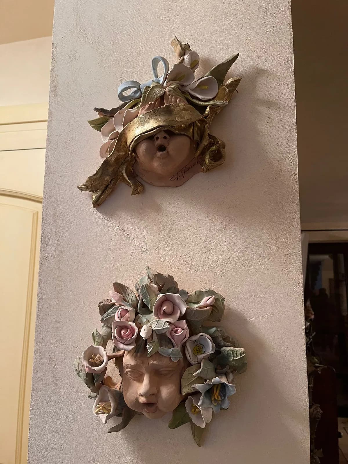Una maschera di ceramica applicata alla parete, con una corona di rose.