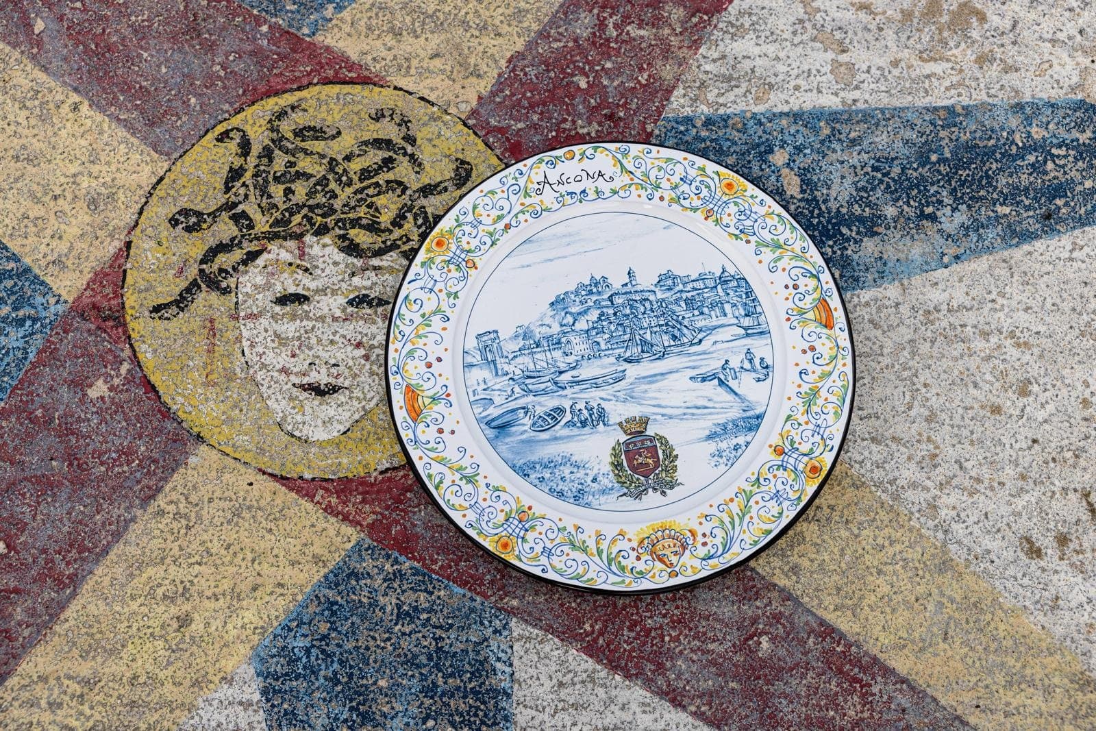 Maiolica plate dedicated to Ancona resting on an ancient mosaic floor with a face and geometric motifs.