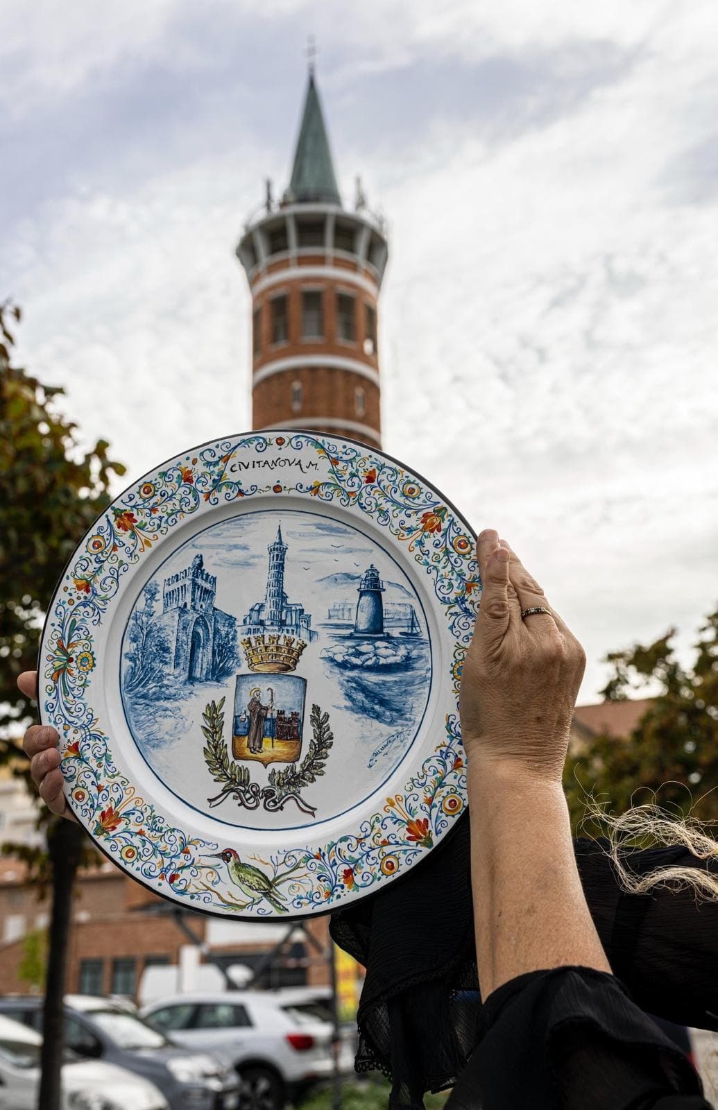 A hand raises a maiolica plate towards the sky in front of the civic tower.