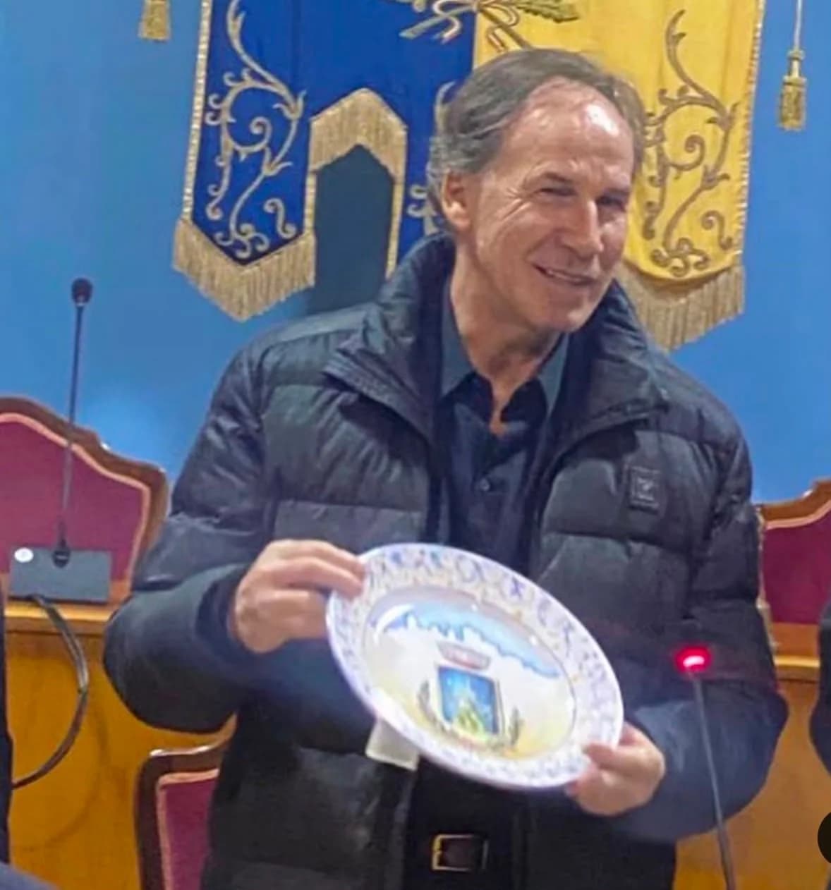In una sala consiliare, un uomo mostra al pubblico il piatto ricevuto.