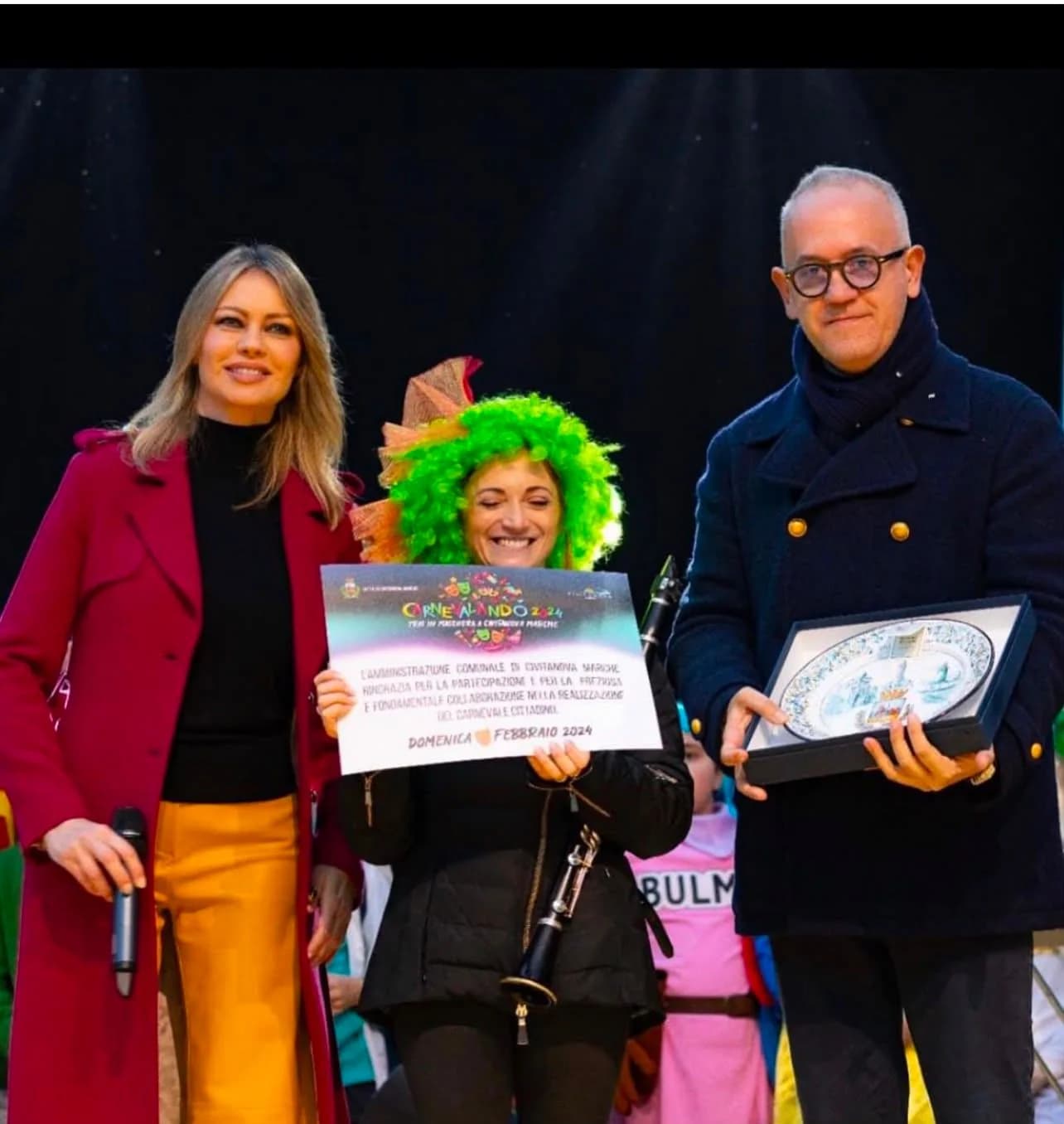 Premiazione di carnevale: tre persone sul palco, una regge il piatto incorniciato.