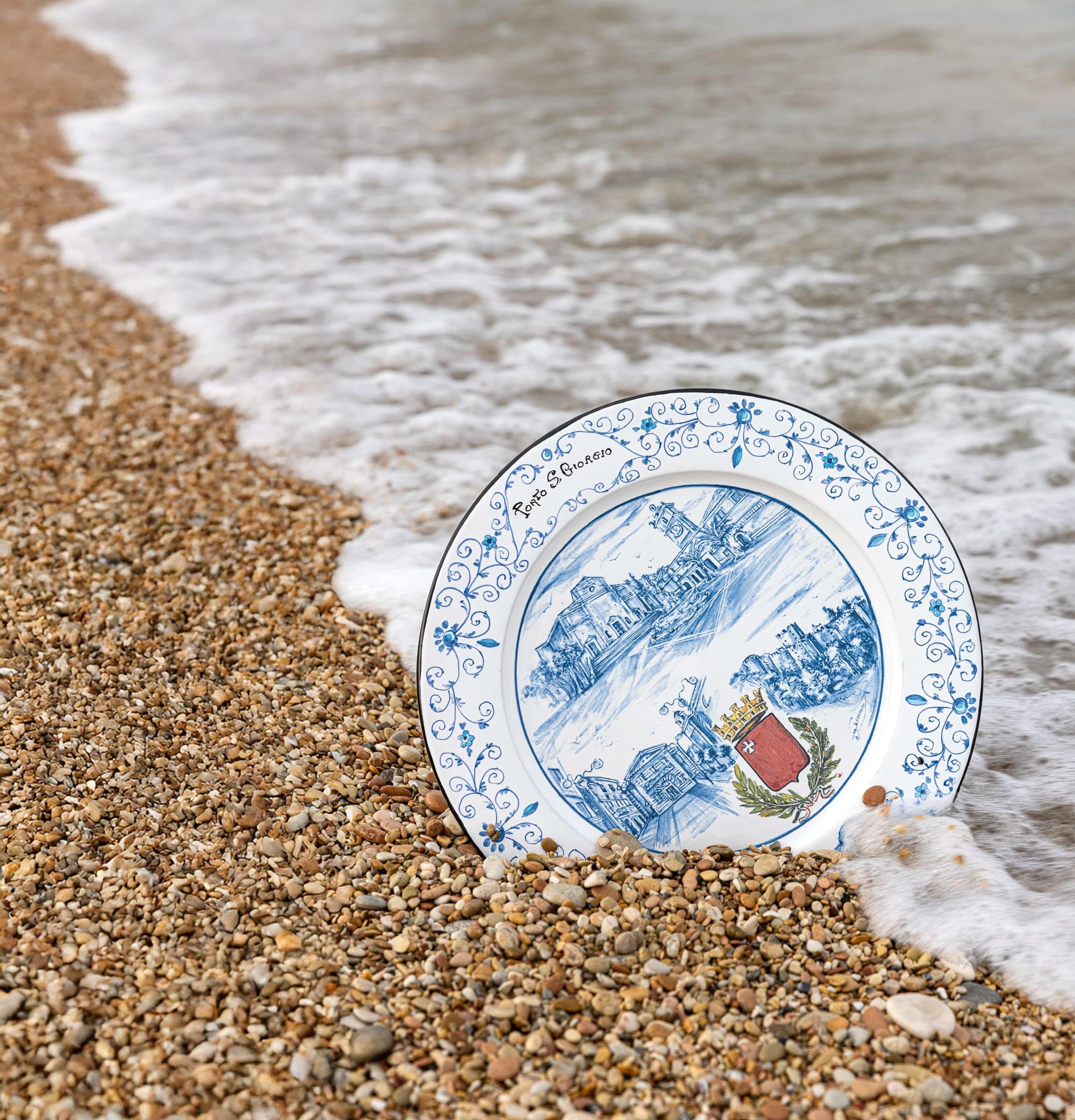 Piatto in maiolica dedicato a Porto San Giorgio, poggiato sui ciottoli della battigia mentre la schiuma dell'onda lo raggiunge.