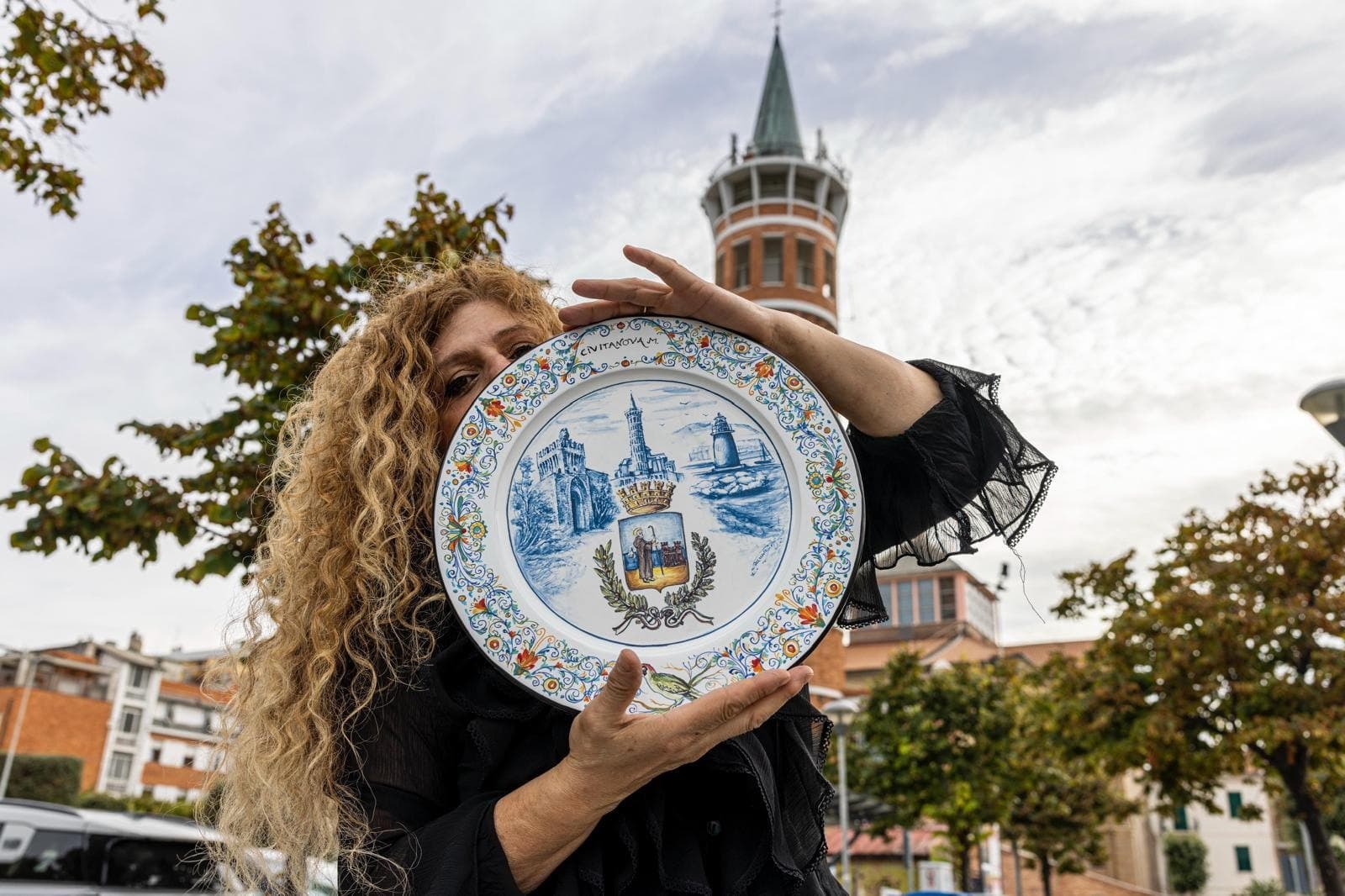 Paola Iannucci solleva con entrambe le mani un piatto in maiolica davanti alla torre civica di Montegranaro.