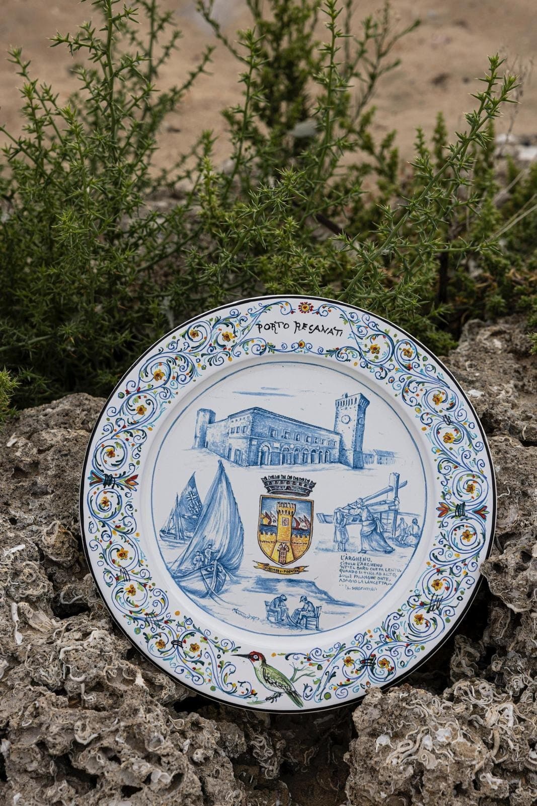 Piatto in maiolica dedicato a Porto Recanati, appoggiato fra le rocce e la vegetazione della scogliera.